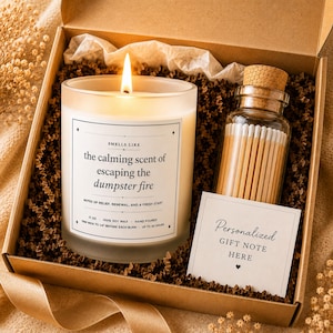 May include: A gift box containing a lit candle in a frosted glass jar with a label reading "SMELLS LIKE the calming scent of escaping the dumpster fire", a glass bottle filled with wooden matches, and a card labeled "Personalized GIFT NOTE HERE".
