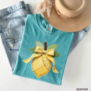 May include: A seafoam green t-shirt featuring a lemon applique with a yellow bow and green leaves. The lemon design incorporates floral and gingham patterns. The shirt is paired with light wash jeans and a tan hat.