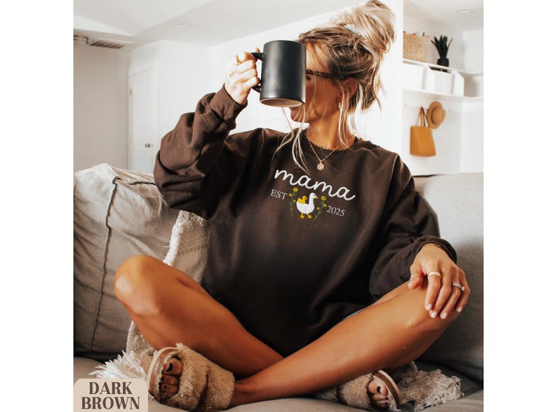 May include: A woman wearing a brown sweatshirt with the text "mama est 2025" and a white duck graphic. She is sitting on a couch and holding a black mug.