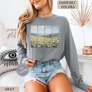 May include: Gray long-sleeve sweatshirt featuring a landscape print of a field of white flowers and a blue sky. The words "COMFORT COLORS" and "FAST SHIPPING" are printed on the image. The word "GRAY" is printed at the bottom.