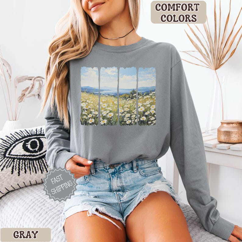 May include: Gray long-sleeve sweatshirt featuring a landscape print of a field of white flowers and a blue sky. The words "COMFORT COLORS" and "FAST SHIPPING" are printed on the image. The word "GRAY" is printed at the bottom.