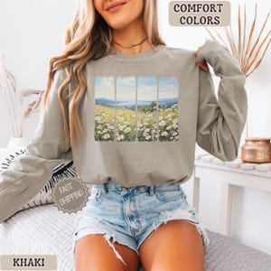 May include: Khaki-colored long-sleeve sweatshirt with a landscape design. The design features a field of white flowers, a blue sky, and a mountain range. The words "COMFORT COLORS" and "KHAKI" are printed on the image.