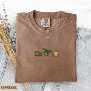 May include: Espresso-colored t-shirt with a crew neck and embroidered green frogs. The shirt features the Comfort Colors brand label and is paired with a silver necklace. The shirt is displayed on a denim background.