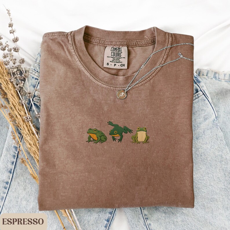 May include: Espresso-colored t-shirt with a crew neck and embroidered green frogs. The shirt features the Comfort Colors brand label and is paired with a silver necklace. The shirt is displayed on a denim background.