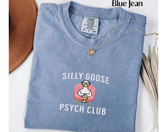 May include: A light blue t-shirt with a white graphic of a goose wearing glasses and sitting in a pink armchair. The text "Silly Goose Psych Club" is printed below the graphic.