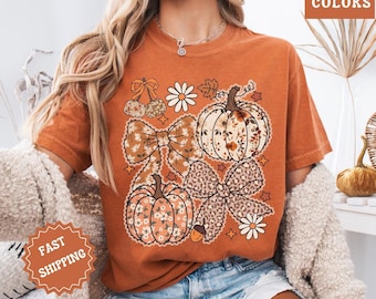 May include: Burnt orange t-shirt featuring a fall-themed graphic. The design includes pumpkins, bows, cherries, and daisies. The words "COMFORT COLORS" are printed on the upper right corner. The shirt has a relaxed fit and short sleeves.