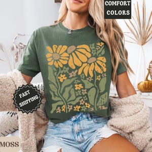 May include: A moss green t-shirt featuring a graphic print of yellow and light green flowers with stems and leaves. The shirt has a crew neckline and short sleeves. Text on the image includes COMFORT COLORS, FAST SHIPPING, and MOSS.