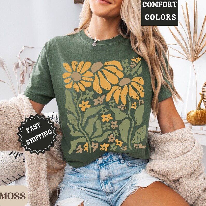 May include: A moss green t-shirt featuring a graphic print of yellow and light green flowers with stems and leaves. The shirt has a crew neckline and short sleeves. Text on the image includes COMFORT COLORS, FAST SHIPPING, and MOSS.