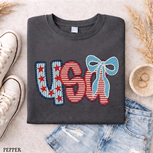 Op de afbeelding: Donkergrijze sweatshirt met het woord "USA" in rood, wit en blauw applicatie. De "U" heeft rode sterren op een blauwe achtergrond, de "S" en "A" hebben rood-witte strepen en een blauwe strik. Witte sneakers en een spijkerbroekshort staan in de afbeelding.