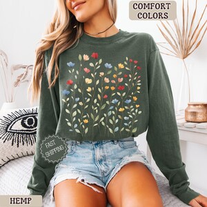 May include: A dark green sweatshirt with a floral design featuring red, blue, and yellow flowers. The words "COMFORT COLORS" and "HEMP" are visible. The sweatshirt is paired with denim shorts.