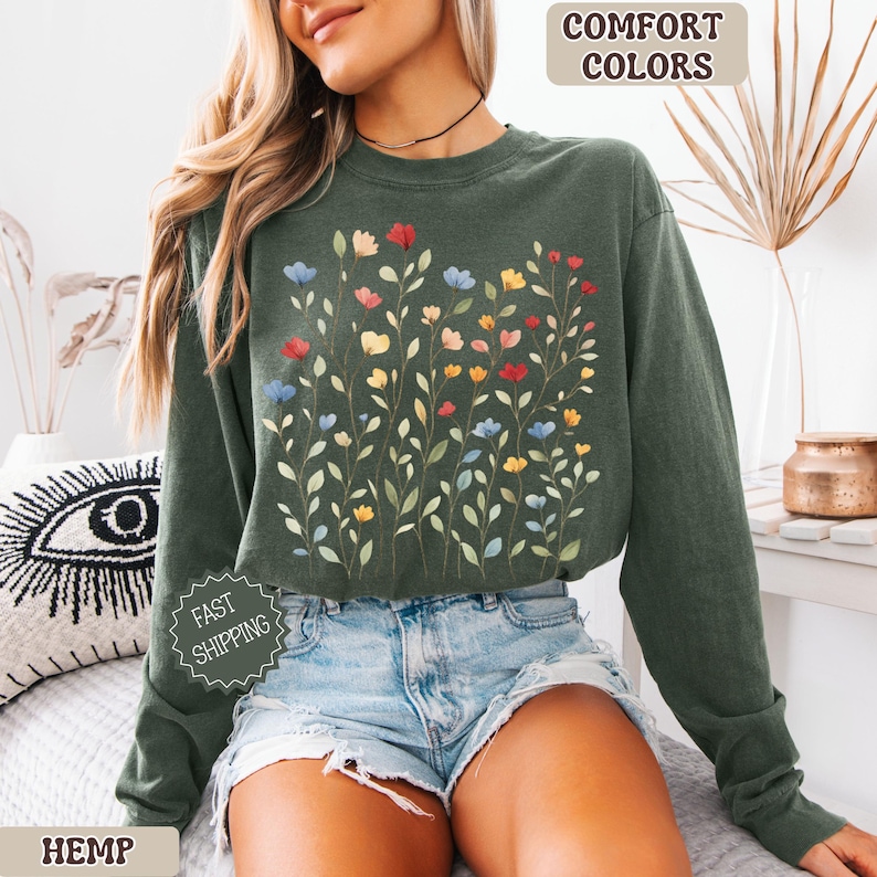 May include: A dark green sweatshirt with a floral design featuring red, blue, and yellow flowers. The words "COMFORT COLORS" and "HEMP" are visible. The sweatshirt is paired with denim shorts.