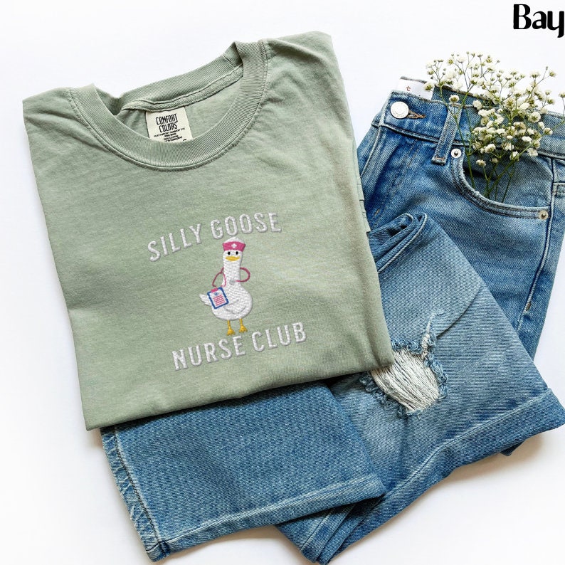 Puede incluir: Camiseta verde salvia con el texto bordado "SILLY GOOSE NURSE CLUB" y un dibujo de un ganso con gorro de enfermera y sujetando una carpeta. La camiseta se combina con vaqueros azules con una rodilla desgastada y un peque&ntilde;o ramo de flores blancas.