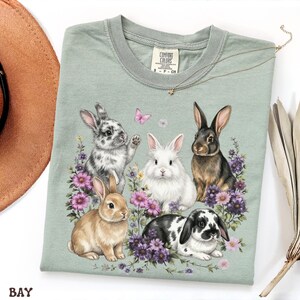 Puede incluir: Una camiseta Comfort Colors verde claro con un gr&aacute;fico de cinco conejos rodeados de flores moradas y una mariposa rosa. Los conejos son de varios colores, incluyendo blanco, marr&oacute;n y negro. Tambi&eacute;n se ve un collar dorado con un colgante de estrella.