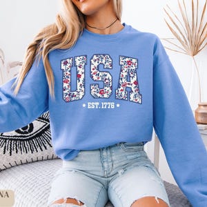 Peut inclure: Un sweat-shirt bleu clair avec le mot "USA" dans un motif floral. Le texte "EST. 1776" est en dessous du mot "USA". Le sweat-shirt est de style col rond.