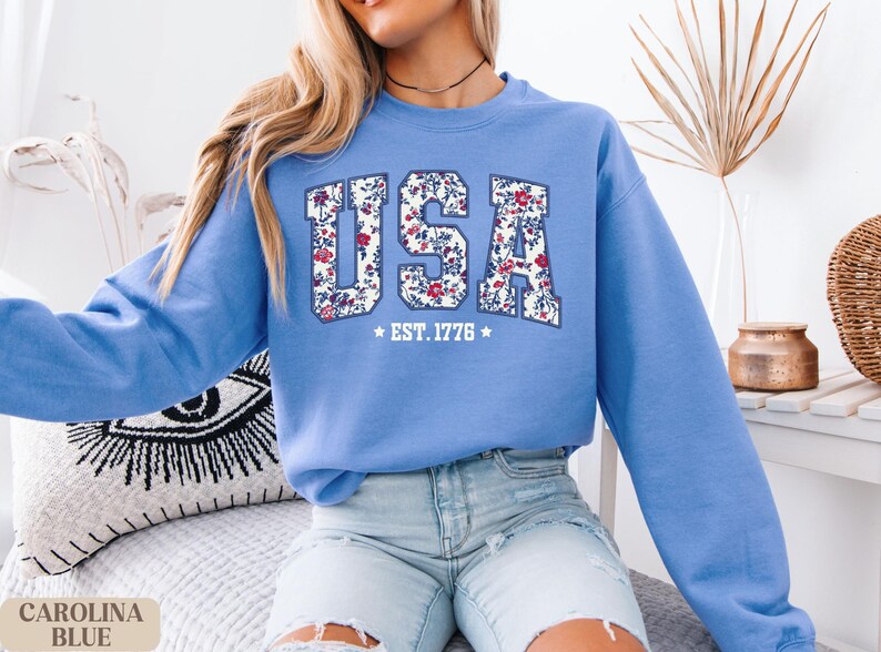 Peut inclure: Un sweat-shirt bleu clair avec le mot "USA" dans un motif floral. Le texte "EST. 1776" est en dessous du mot "USA". Le sweat-shirt est de style col rond.