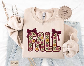 May include: A sand-colored sweatshirt with the word "FALL" in a floral design. The letters are outlined in a dark burgundy color and decorated with burgundy bows. The sweatshirt has a "Super Soft" tag.