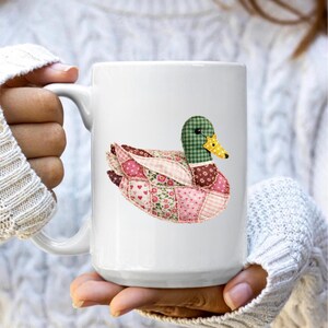 May include: White ceramic mug featuring a patchwork duck design. The duck has a green checkered head, a yellow beak, and a body composed of pink and red floral and heart-patterned fabric. The mug is held by a person wearing a white sweater.