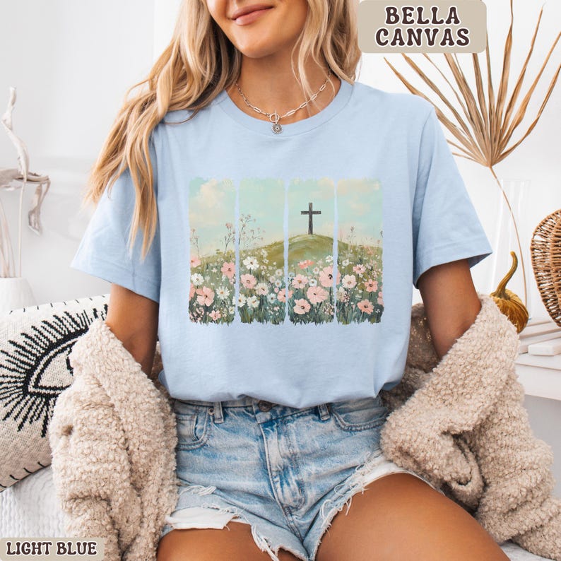 May include: Light blue Bella Canvas t-shirt featuring a floral and cross design. The graphic shows a cross on a hill with flowers. The shirt is light blue and has the words "Bella Canvas" printed on the top right corner. The words "Light Blue" are printed on the bottom left corner.