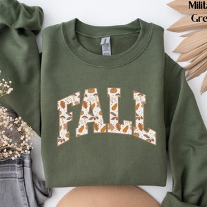 K&ouml;nnte beinhalten: Ein gr&uuml;ner Pullover mit dem Wort "FALL" in wei&szlig;en Buchstaben mit einem Herbstlaubmuster. Der Pullover ist ein Rundhalsmodell.