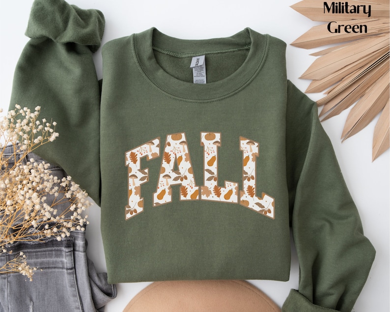 K&ouml;nnte beinhalten: Ein gr&uuml;ner Pullover mit dem Wort "FALL" in wei&szlig;en Buchstaben mit einem Herbstlaubmuster. Der Pullover ist ein Rundhalsmodell.
