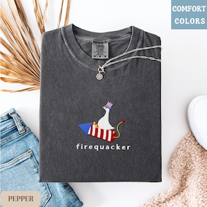 K&ouml;nnte beinhalten: Dunkelgraues T-Shirt mit einem gestickten Design einer Gans auf einer Rakete, die einen patriotischen Hut tr&auml;gt. Das Wort "firequacker" ist darunter gestickt. Eine silberne Halskette und eine Jeans liegen neben dem Shirt.