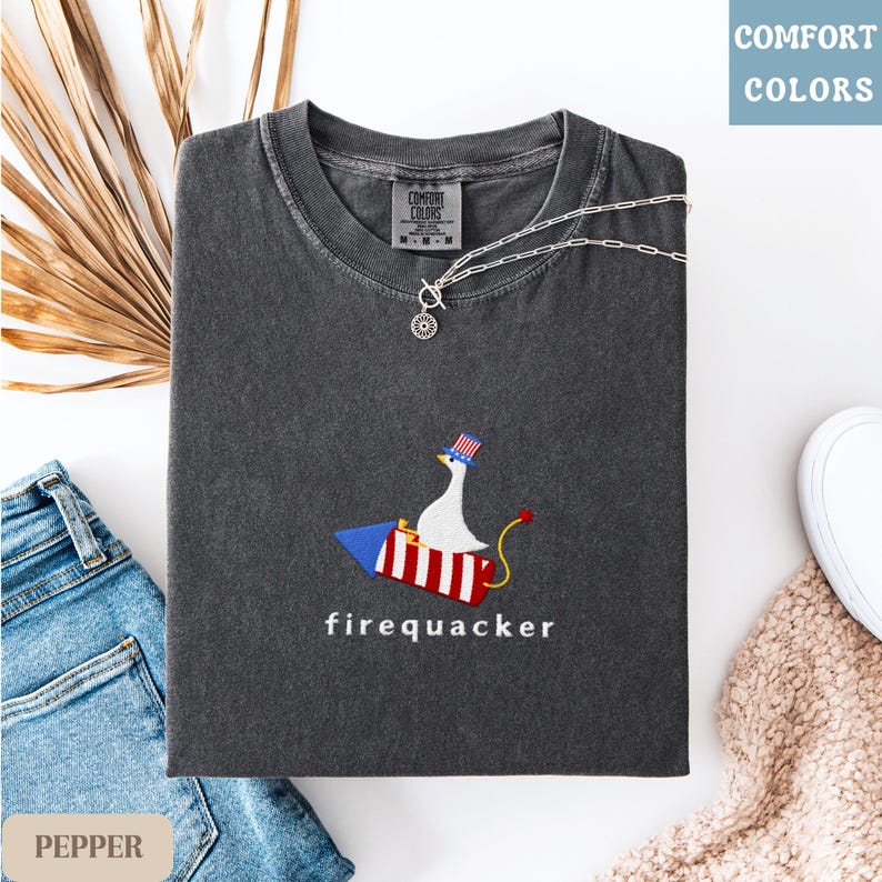 K&ouml;nnte beinhalten: Dunkelgraues T-Shirt mit einem gestickten Design einer Gans auf einer Rakete, die einen patriotischen Hut tr&auml;gt. Das Wort "firequacker" ist darunter gestickt. Eine silberne Halskette und eine Jeans liegen neben dem Shirt.