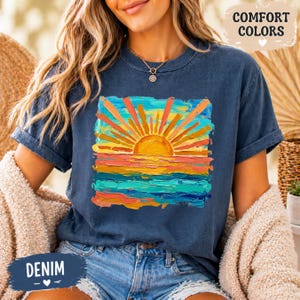 Puede incluir: Camiseta azul denim con un gr&aacute;fico de puesta de sol colorido. La obra de arte presenta un sol vibrante con rayos naranjas y amarillos sobre un mar de pinceladas azules, turquesas y naranjas. Las palabras "COMFORT COLORS" y "DENIM" est&aacute;n impresas.