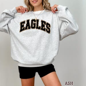 May include: A light gray sweatshirt with the word "EAGLES" in a black and tan leopard print design across the chest. The sweatshirt has a crew neck and long sleeves. The model is wearing black shorts.