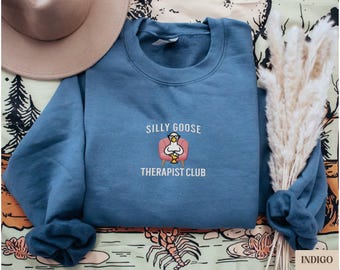 May include: A blue sweatshirt with the text "Silly Goose Therapist Club" and an embroidered image of a goose wearing glasses and sitting in a pink armchair.