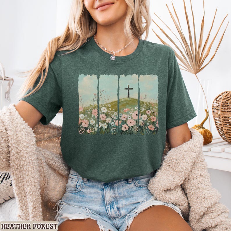 May include: Heather forest green t-shirt featuring a graphic of a cross on a hill, surrounded by wildflowers. The design is framed by vertical lines. The shirt is a crew neck and short-sleeved.