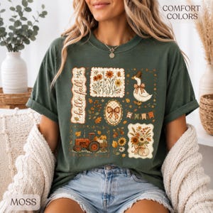 May include: A dark green short-sleeve t-shirt featuring a graphic print with the text "hello fall" and "COMFORT COLORS". The design includes a goose wearing a hat, a tractor, floral arrangements, a bow, and autumn leaves in orange and brown.