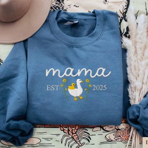 May include: Blue sweatshirt with white embroidery that reads "mama est 2025" and features a white duck with a yellow duckling.