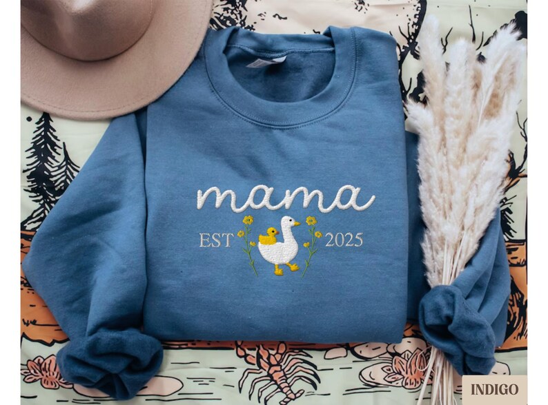 May include: Blue sweatshirt with white embroidery that reads "mama est 2025" and features a white duck with a yellow duckling.