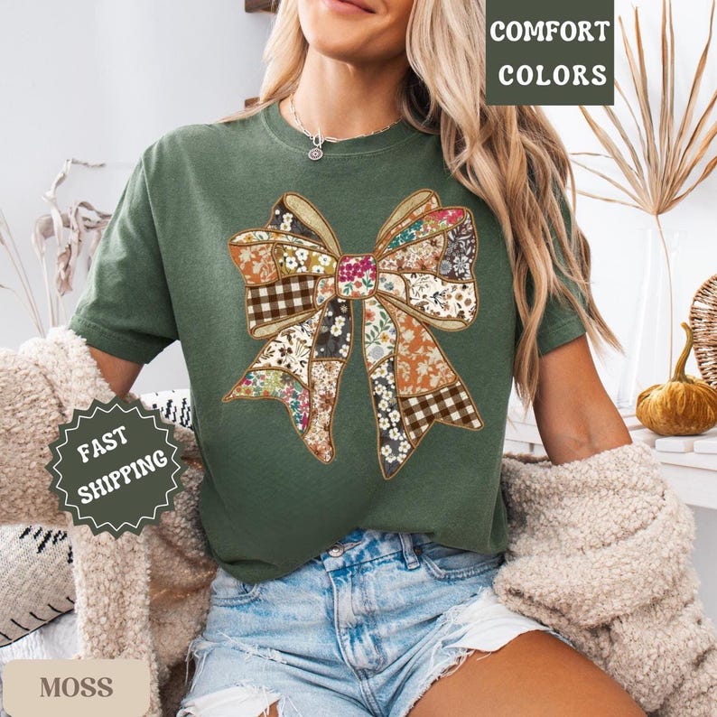 May include: A moss green t-shirt featuring a patchwork bow design with floral and plaid patterns. The shirt has short sleeves and a crew neck. The words "COMFORT COLORS" and "MOSS" are visible. The shirt is part of a clothing collection.