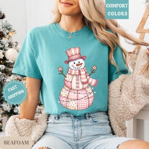 May include: Seafoam green t-shirt featuring a whimsical snowman design. The snowman wears a pink top hat, scarf, and is adorned with star-shaped arms. The shirt has the words "COMFORT COLORS" and "FAST SHIPPING" printed on it.