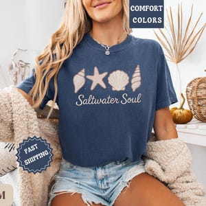 May include: A navy blue t-shirt with the words "Saltwater Soul" and seashell graphics. The shirt has a crew neck and short sleeves. The shirt is made of a soft, comfortable material.