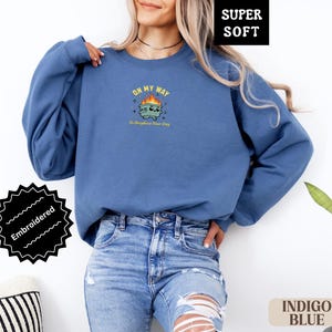 Pu&ograve; includere: Felpa blu indaco con grafica ricamata. La grafica presenta un bidone della spazzatura dei cartoni animati con fiamme e il testo "ON MY WAY To Brighten Your Day". La felpa &egrave; etichettata "SUPER SOFT" e "Embroidered".