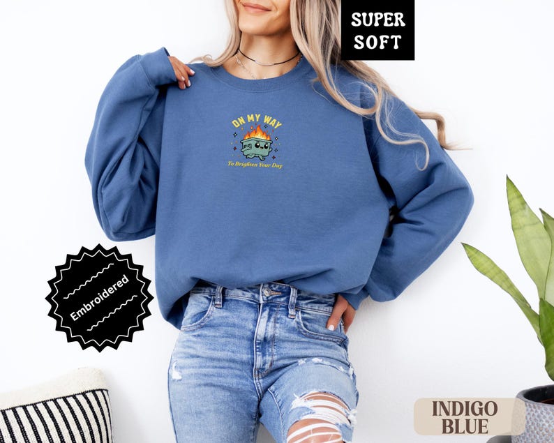 Pu&ograve; includere: Felpa blu indaco con grafica ricamata. La grafica presenta un bidone della spazzatura dei cartoni animati con fiamme e il testo "ON MY WAY To Brighten Your Day". La felpa &egrave; etichettata "SUPER SOFT" e "Embroidered".