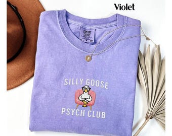 May include: A purple t-shirt with the text "Silly Goose Psych Club" and a cartoon goose wearing glasses sitting in a pink armchair.