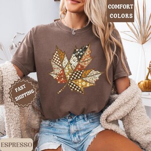 May include: Espresso-colored t-shirt featuring a large, patchwork-style maple leaf design. The leaf incorporates various patterns and colors, including floral, gingham, and solid patches. The shirt has short sleeves and a relaxed fit. The words "Comfort Colors" are printed on the top right.