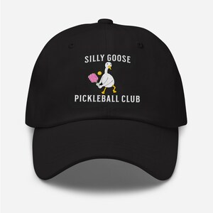 May include: Black baseball cap with white embroidery that reads "Silly Goose Pickleball Club". A white goose with yellow feet is holding a pink and white pickleball paddle and a yellow ball.