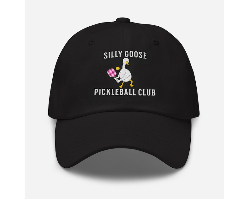 May include: Black baseball cap with white embroidery that reads "Silly Goose Pickleball Club". A white goose with yellow feet is holding a pink and white pickleball paddle and a yellow ball.