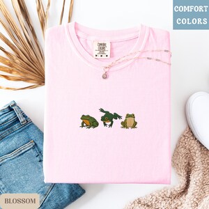 May include: A light pink t-shirt with three embroidered frogs in green and brown. The shirt has a crew neck and short sleeves. A silver necklace with a small pendant rests on the shirt. The words "COMFORT COLORS" are printed on the shirt's tag.