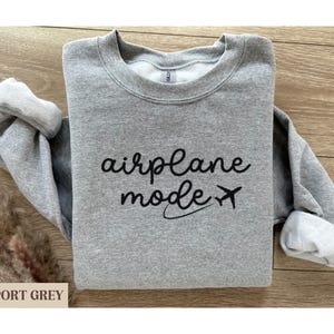 Puede incluir: Una sudadera gris con el texto "modo avi&oacute;n" y un peque&ntilde;o icono de avi&oacute;n impreso en la parte delantera.