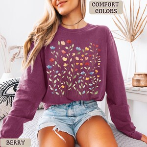 May include: Berry-colored sweatshirt with a floral design. The design features colorful flowers in red, blue, yellow, and orange on a stem with green leaves. The words "COMFORT COLORS" and "BERRY" are printed on the image.