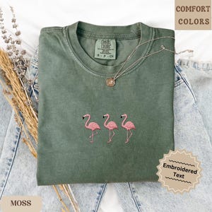 May include: A moss green Comfort Colors t-shirt with three embroidered pink flamingos. The shirt has a crew neck and short sleeves. A silver necklace with a pendant rests on the shirt. The words "Embroidered Text" are on a tag.