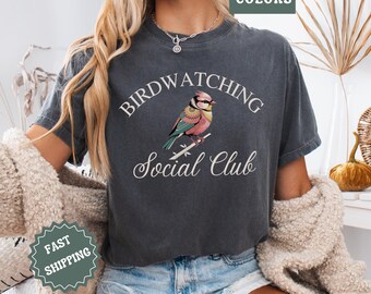 May include: A dark gray t-shirt with a vintage look featuring a colorful bird illustration and the text "Birdwatching Social Club".