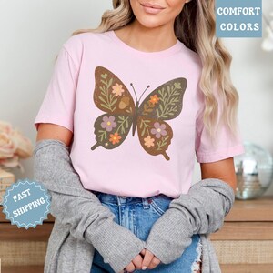 Peut inclure: Un t-shirt rose clair avec un grand motif de papillon. Le papillon est marron avec des d&eacute;tails floraux et de feuilles verts, oranges et violets. Le t-shirt porte le texte "Comfort Colors" dans un encadr&eacute; bleu.