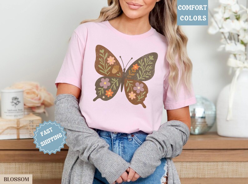 Peut inclure: Un t-shirt rose clair avec un grand motif de papillon. Le papillon est marron avec des d&eacute;tails floraux et de feuilles verts, oranges et violets. Le t-shirt porte le texte "Comfort Colors" dans un encadr&eacute; bleu.