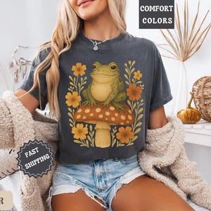 May include: A dark grey t-shirt featuring a graphic of a green frog sitting on a mushroom with yellow flowers. The text "COMFORT COLORS" is in the top right corner of the image.